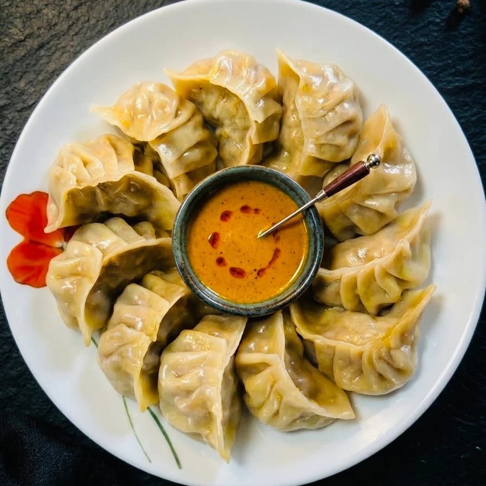 A white plate filled with steamed momos, arranged around a small bowl of orange dipping sauce.